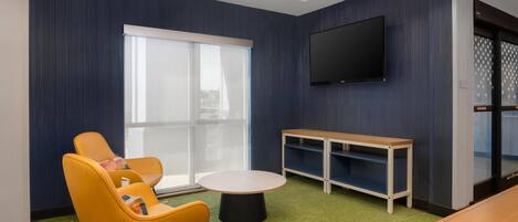 A modern room with a large window, a flat-screen TV, a round table, and a seating area with yellow chairs.