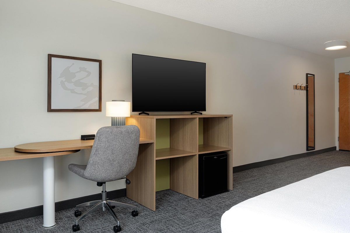 Une chambre d’hôtel comprenant un lit, un bureau avec une chaise, une télévision, un petit réfrigérateur et un tableau encadré au mur.