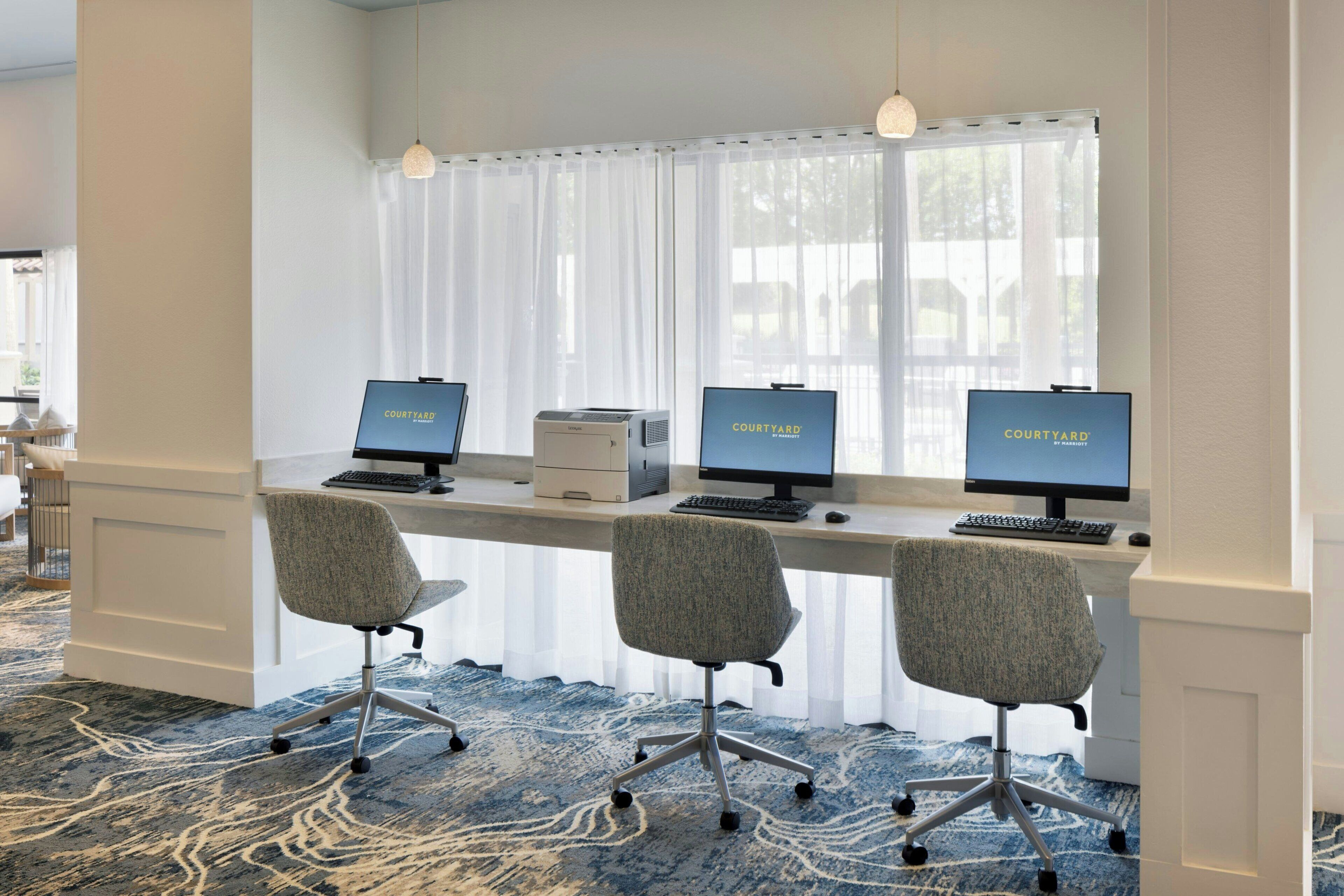 Un espacio de oficina moderno con tres puestos de trabajo equipados con computadoras, cada una mostrando 'COURTYARD'. Hay tres sillas de oficina grises y un escritorio blanco. La habitación tiene un ventanal con cortinas translúcidas, una alfombra estampada e iluminación colgante.