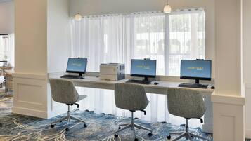 Un espacio de oficina moderno con tres puestos de trabajo equipados con computadoras, cada una mostrando 'COURTYARD'. Hay tres sillas de oficina grises y un escritorio blanco. La habitación tiene un ventanal con cortinas translúcidas, una alfombra estampada e iluminación colgante.