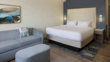 A modern hotel room with a large bed, a grey sofa, a small ottoman, and a painting on the wall.
