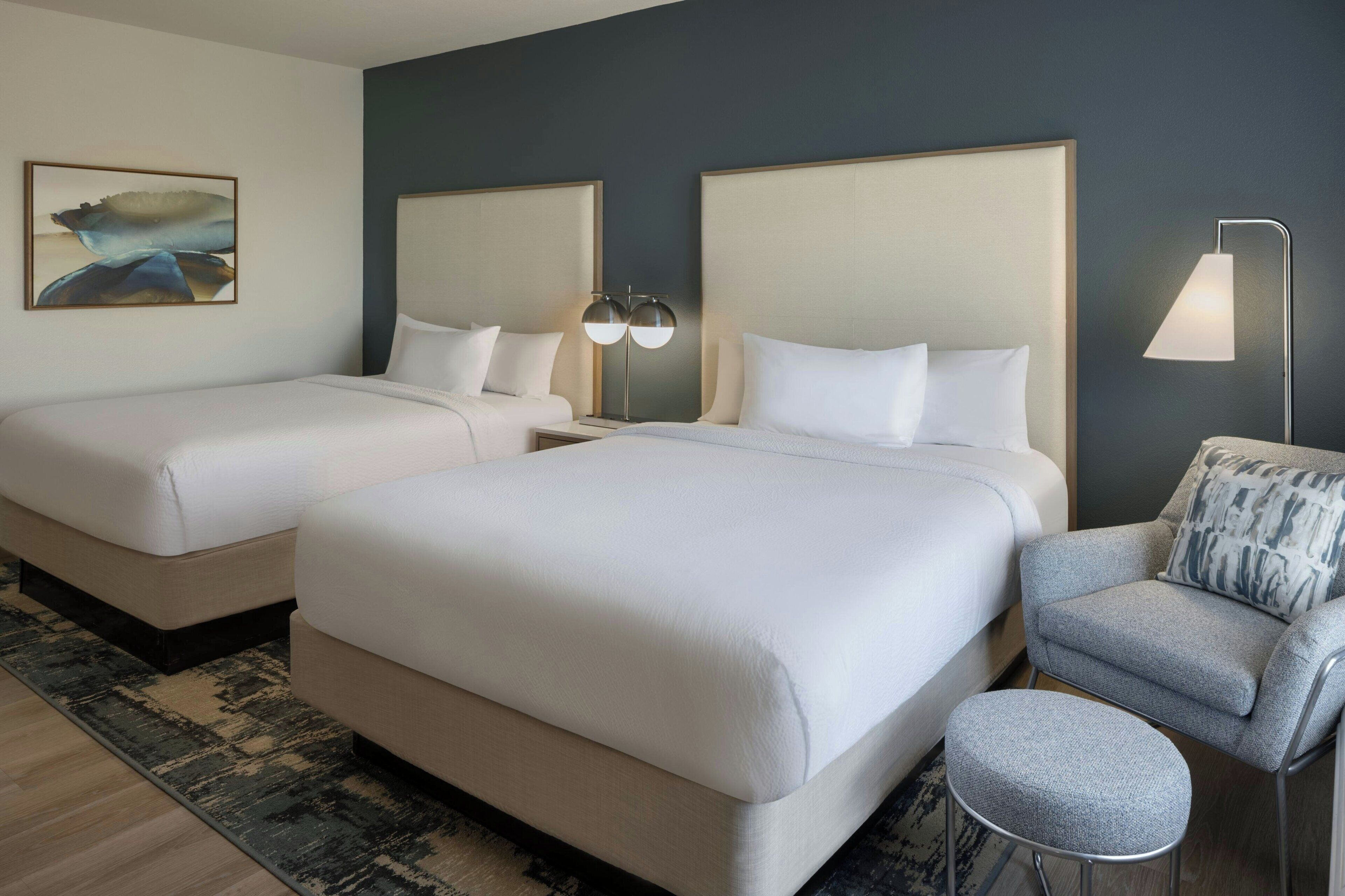 A hotel room with two beds, a grey armchair, a small stool, a nightstand with a lamp, and a painting on the wall.