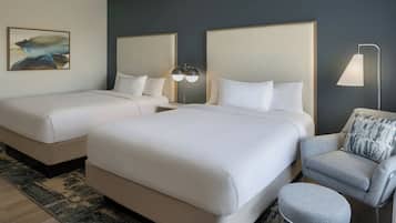 A hotel room with two beds, a grey armchair, a small stool, a nightstand with a lamp, and a painting on the wall.