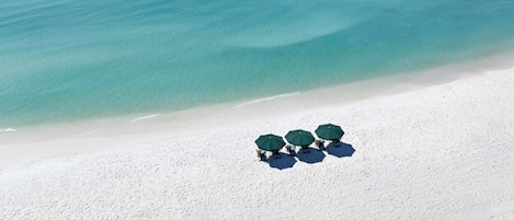 Una playa con arena blanca y mar azul cristalino. Hay tres sombrillas y sillas verdes sobre la arena.