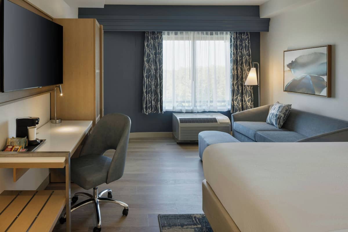 A hotel room with a bed, a desk with a chair, a television, an air conditioning unit, and a window with curtains.