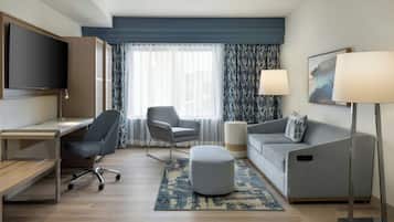 A modern hotel room with a flat-screen TV, a desk, a grey armchair, a grey sofa, a round ottoman, and a floor lamp.