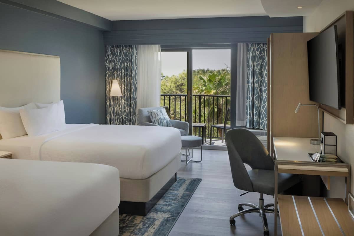 A hotel room with a large bed, a desk, a chair, and a TV. There is a balcony with a view of greenery.