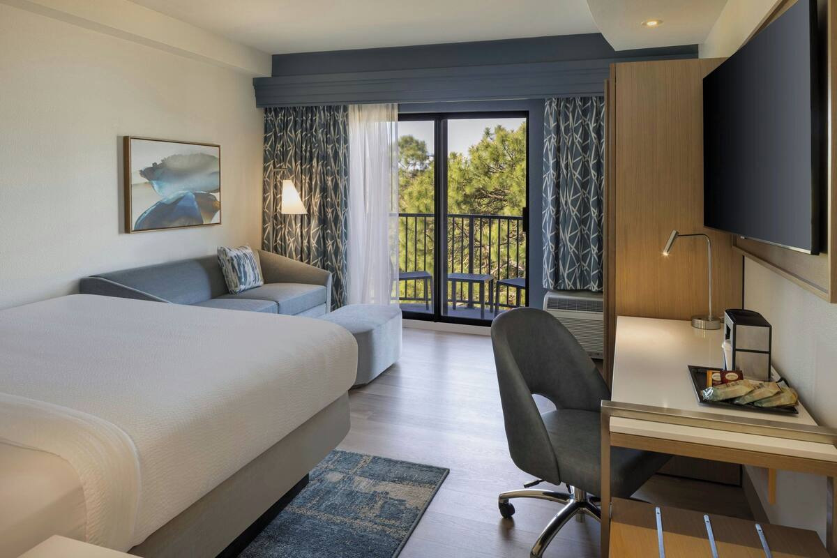 A modern hotel room with a large bed, a desk with a chair, a TV, and a balcony with a view of trees.