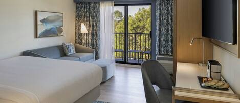 Habitación de hotel moderna con una cama grande, un escritorio con silla, televisor y balcón con vistas a árboles.