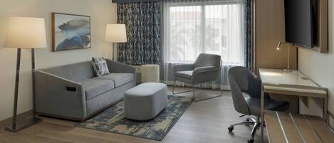 A modern hotel room with a grey sofa, a small ottoman, a desk with a chair, a flat-screen TV, and two floor lamps.