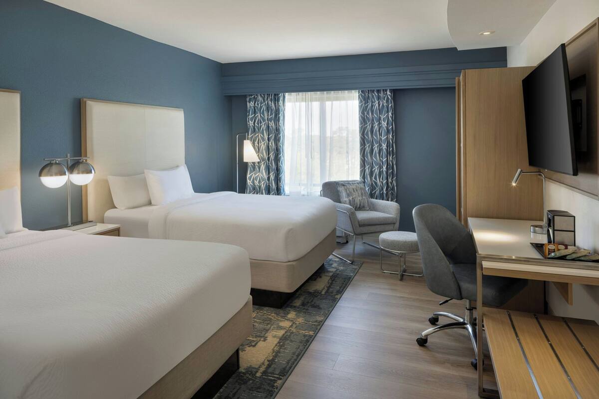 A hotel room with two beds, a desk, a chair, a TV, and a window with curtains.