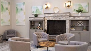 A modern living room with a fireplace, grey upholstered armchairs, a round wooden coffee table, and abstract wall art. - Wyndham Garden Otsego-Minneapolis (Elk River)