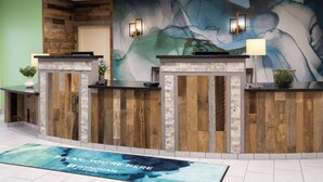 A reception area with wooden reception desks, a large abstract mural, and a welcome mat. - Wyndham Garden Otsego-Minneapolis (Elk River)