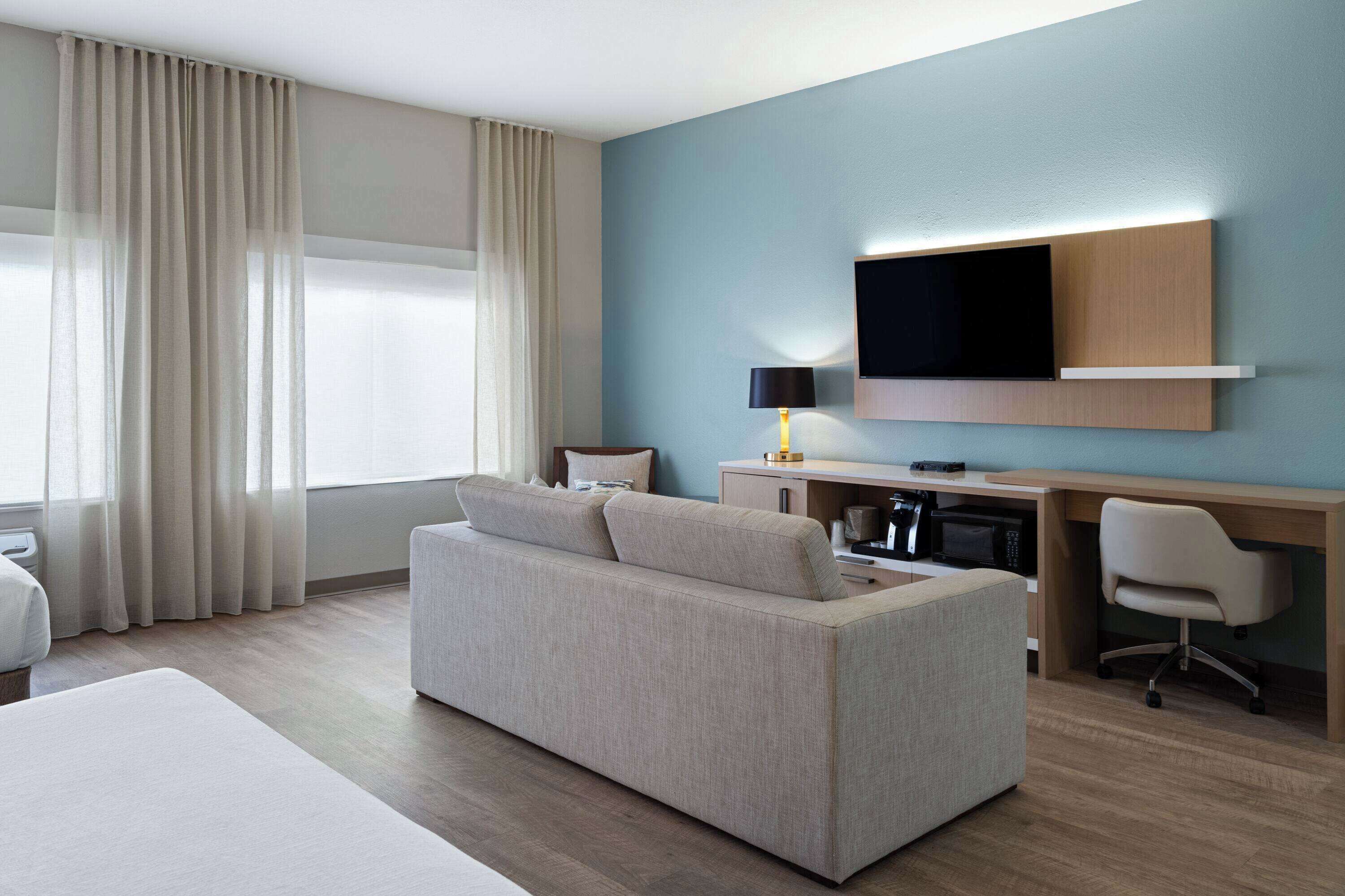 A modern hotel room with a sofa, a desk with a flat-screen TV, a lamp, and a microwave.