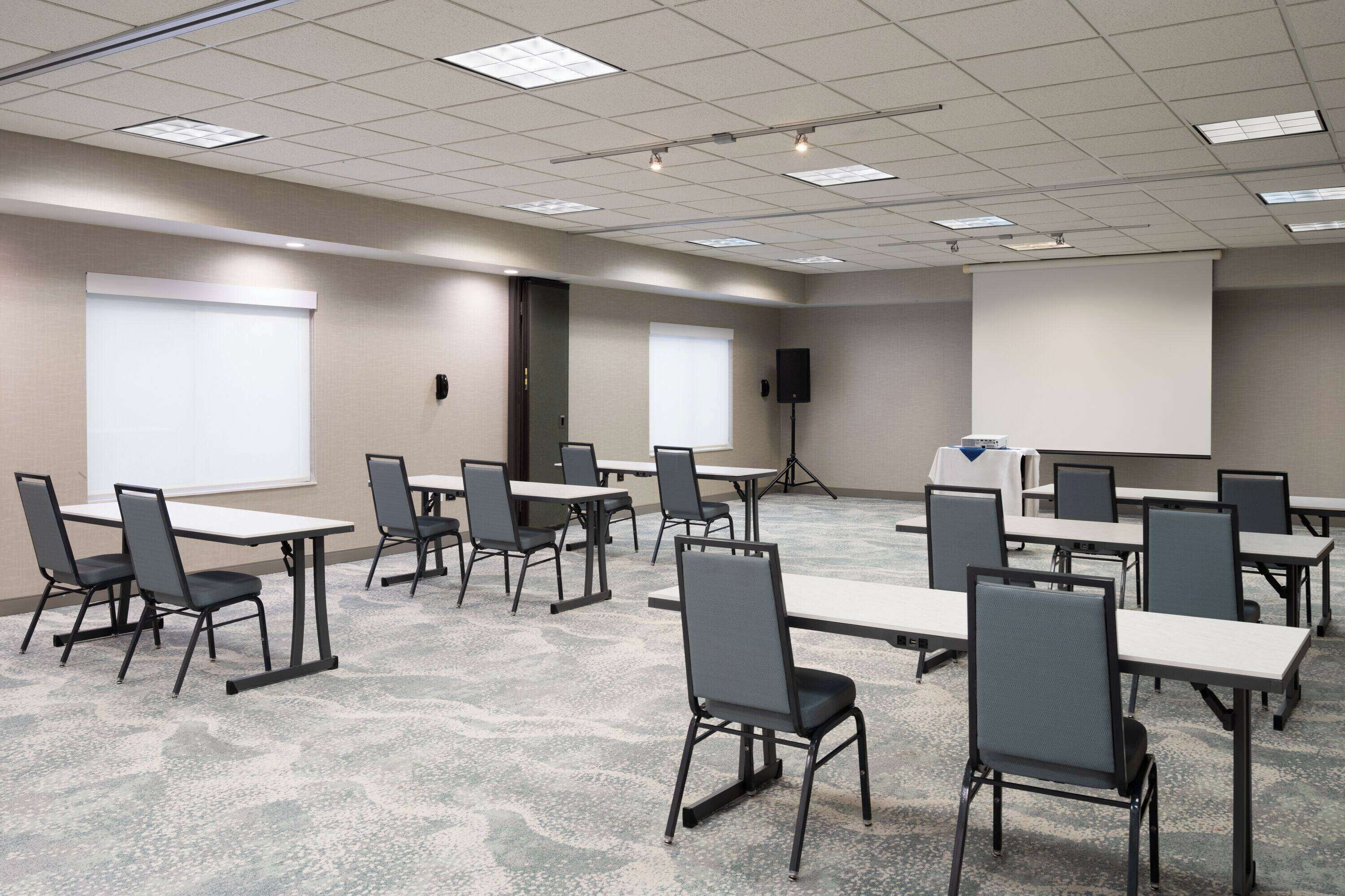 Un salón de conferencias con varias mesas y sillas, equipado con proyectores y una pantalla.