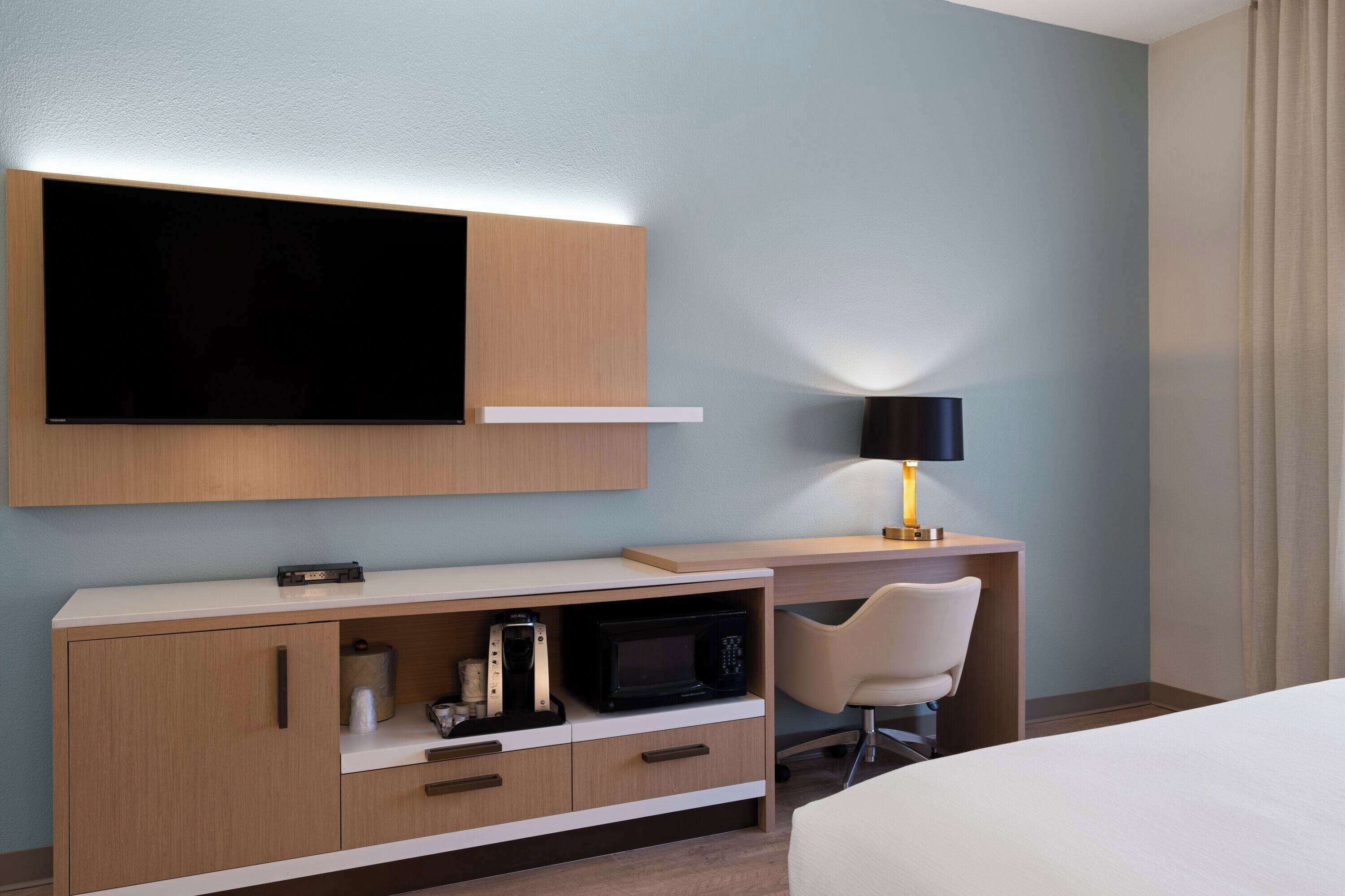 A hotel room with a bed, a desk with a lamp, a microwave, and a TV mounted on the wall.