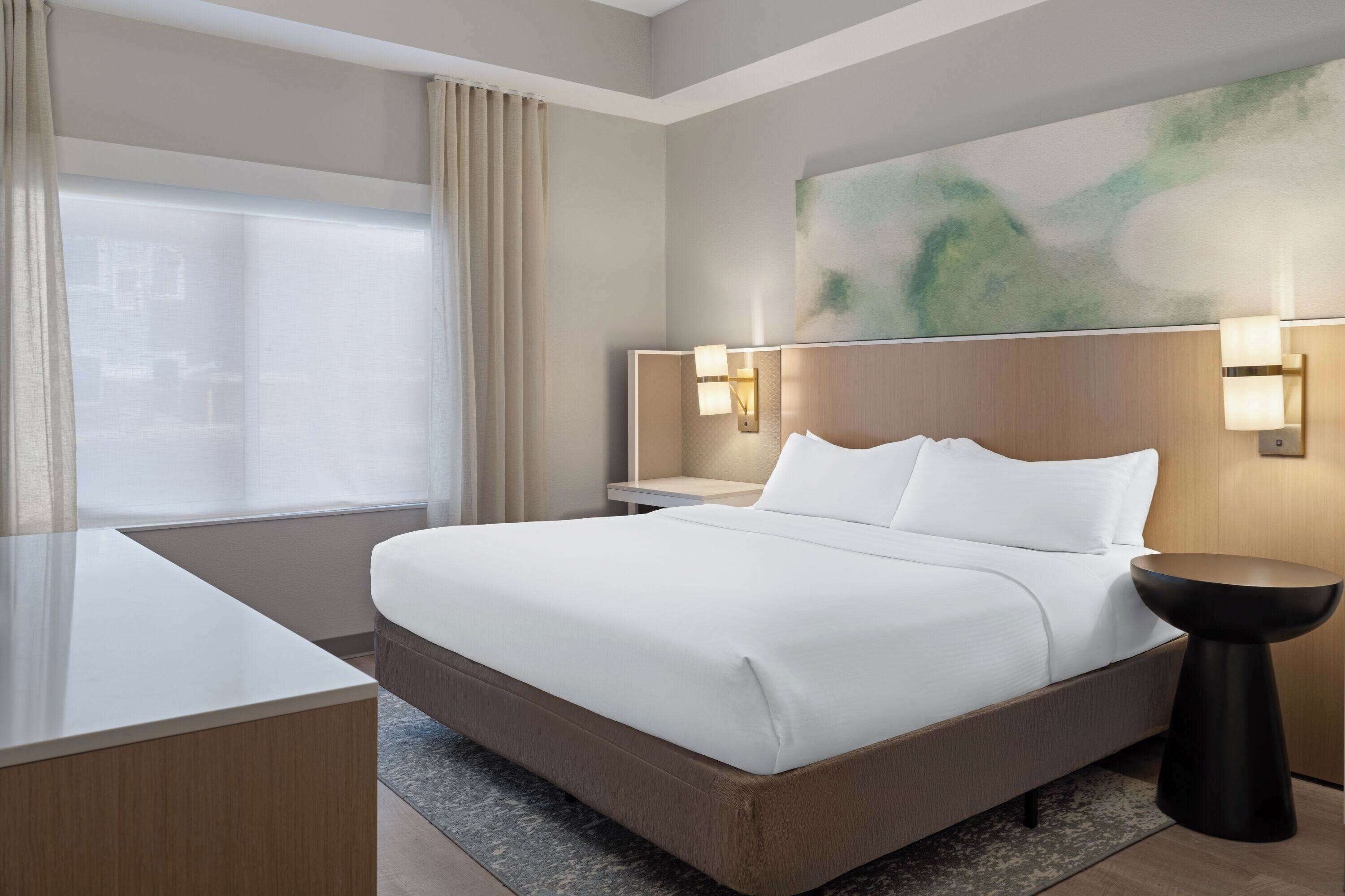 Habitación de hotel con una cama grande, mesitas de noche, un escritorio y vistas a la ciudad por la ventana.