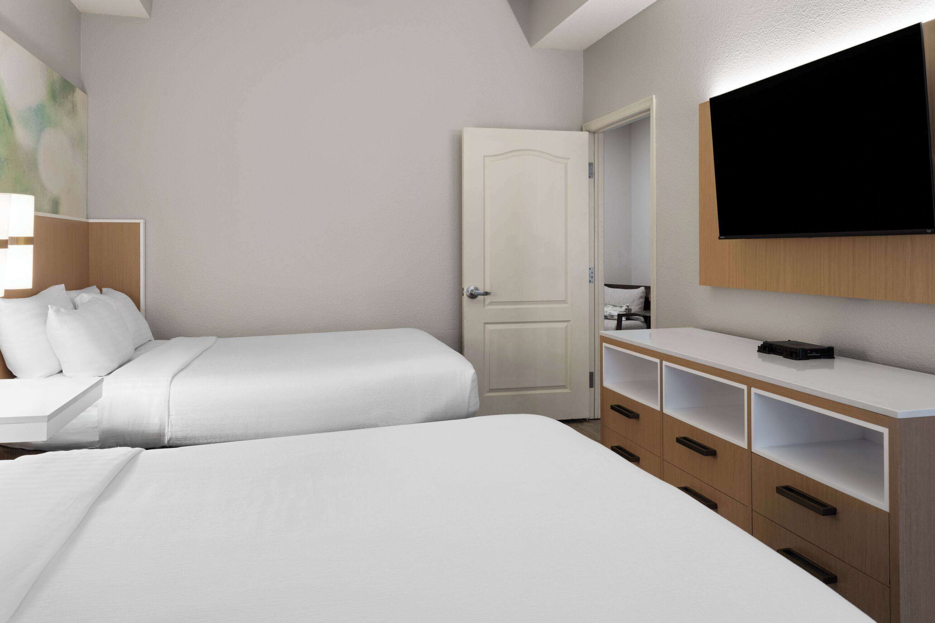 Una habitación de hotel con dos camas, un televisor montado en la pared y un mueble de madera con cajones.