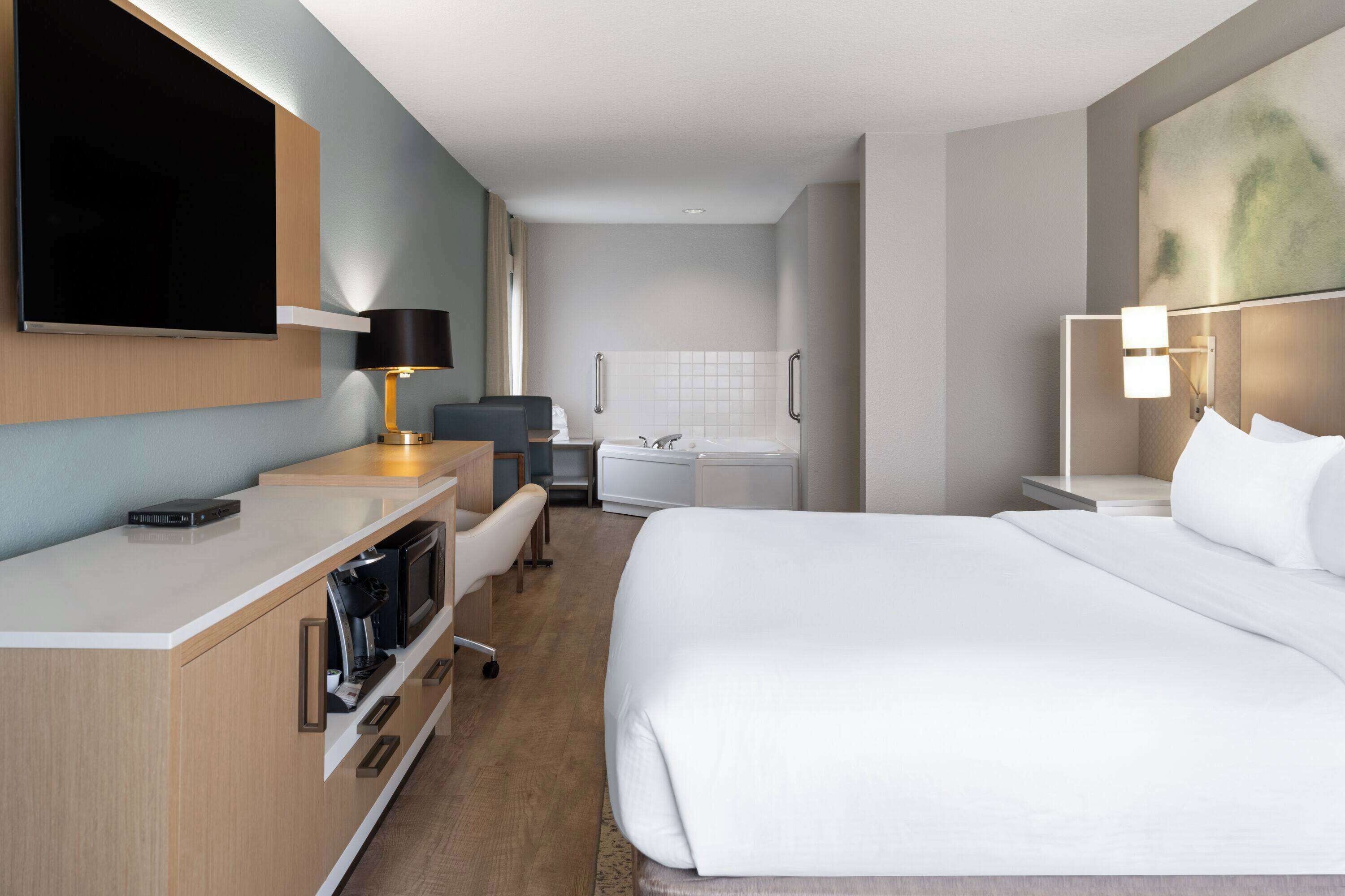 A hotel room with a bed, a desk with a flat-screen TV, a microwave, a coffee maker, a chair, and a bathroom with a bathtub and a sink.