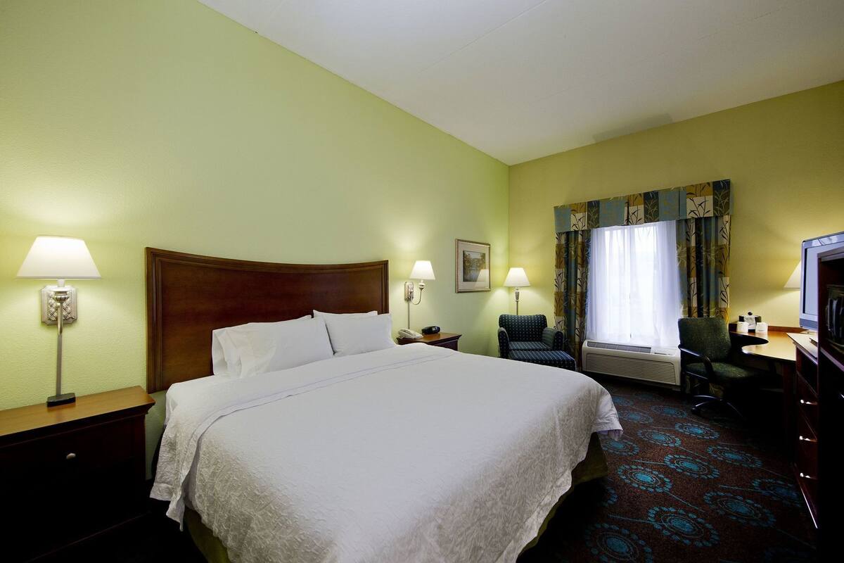 A hotel room with a large bed, two bedside lamps, a chair, a desk with a television, and a window with curtains.