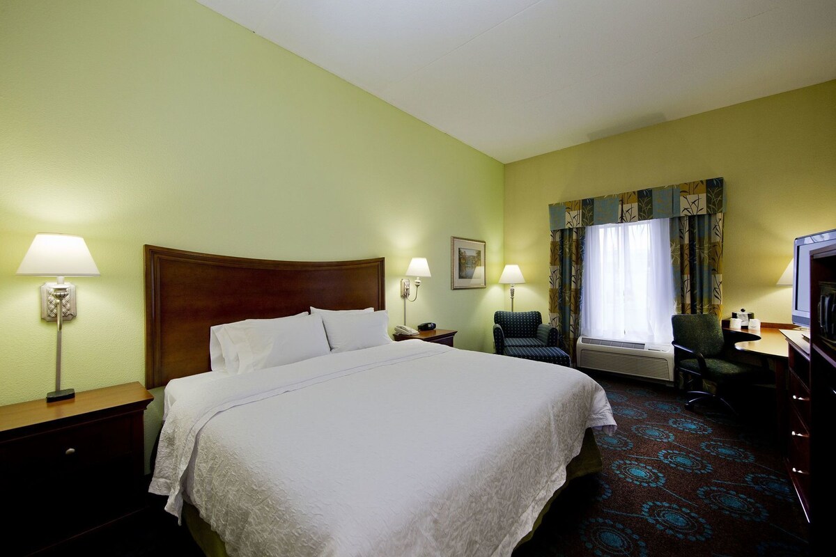 A hotel room with a large bed, two bedside lamps, a chair, a desk with a television, and a window with curtains.