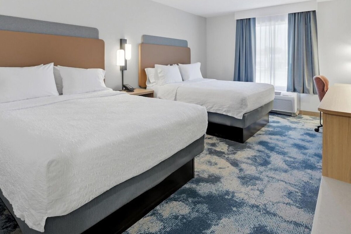 A hotel room with two beds, a desk, a chair, and a window with curtains.