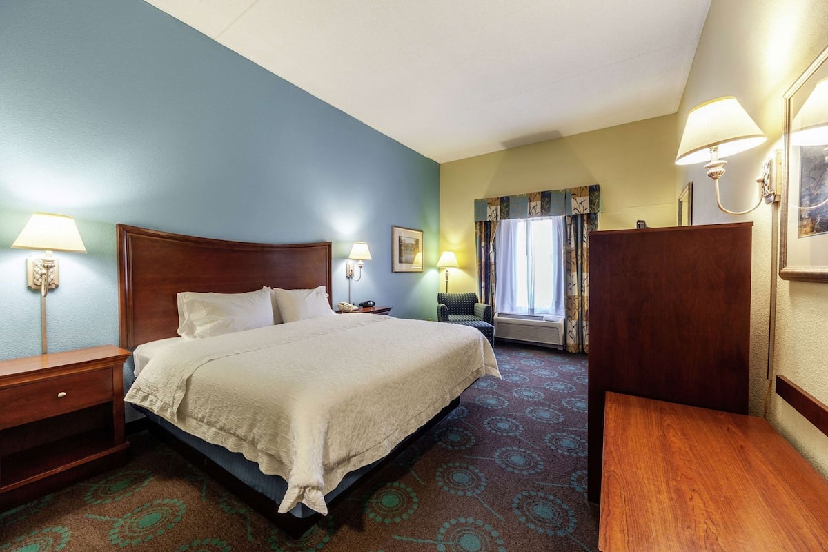 A hotel room with a bed, bedside tables, a chair, a desk, a window with curtains, and wall-mounted lamps.