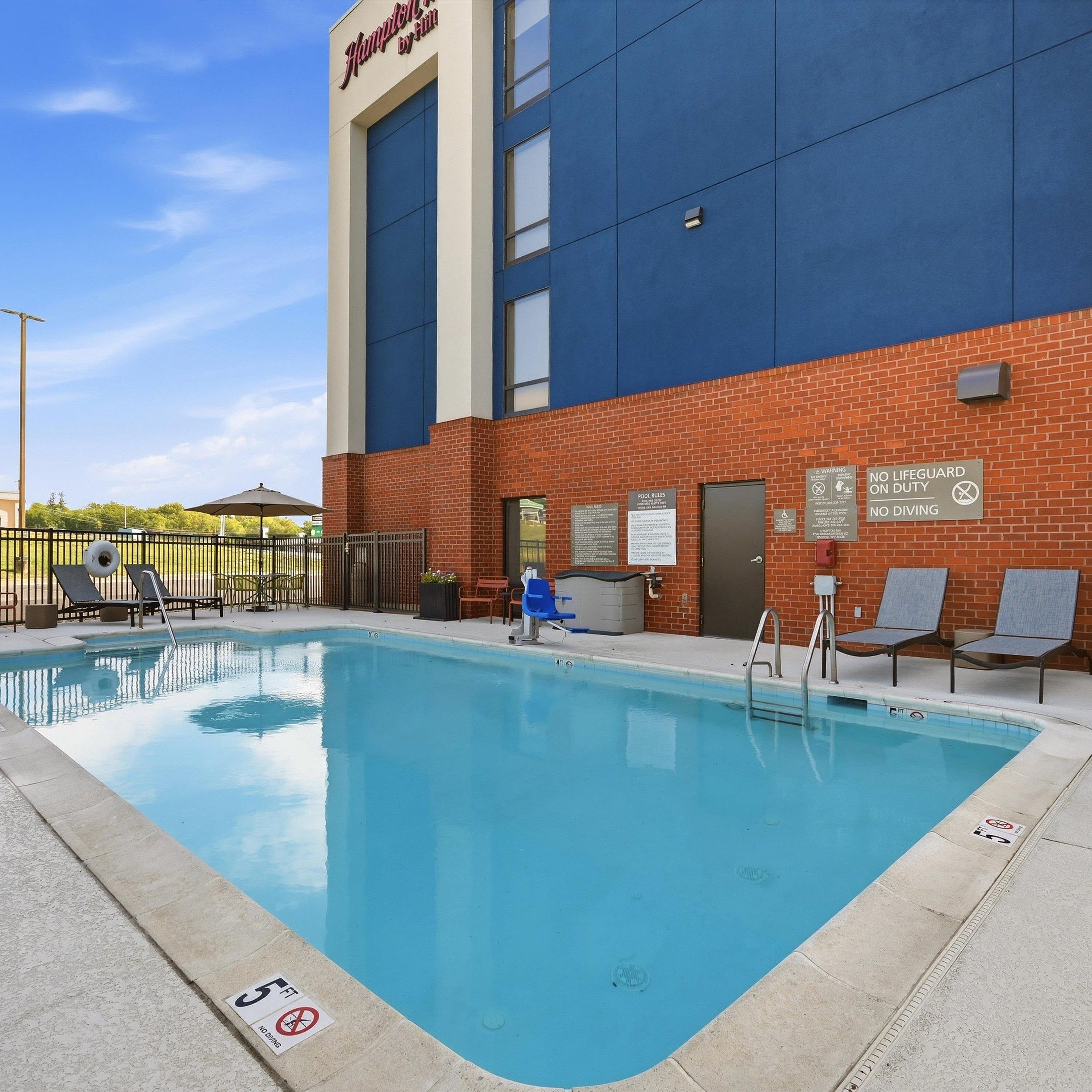 A swimming pool area with lounge chairs and a building with a sign reading 'Hampton by Hilton'.