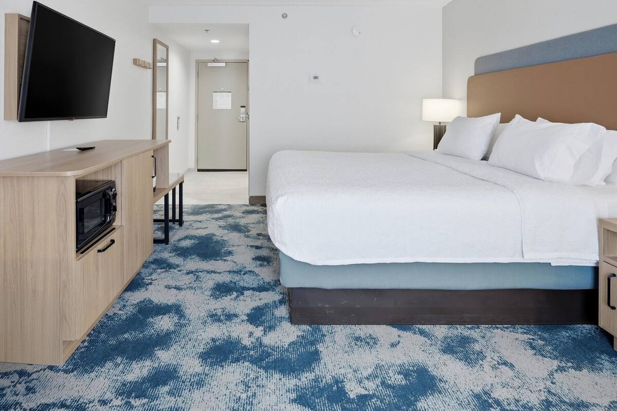 A hotel room with a bed, a TV, a desk with a microwave, and a patterned carpet.