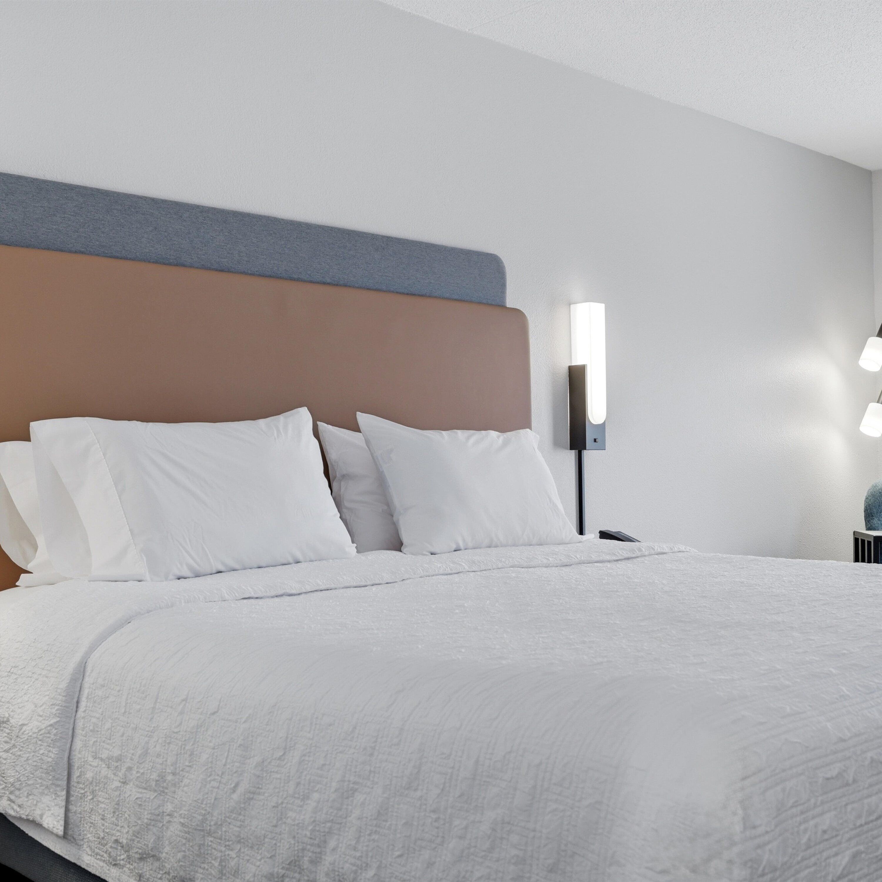 Una cama bien hecha con ropa blanca, un cabecero con panel gris y marrón, y una lámpara de pared.