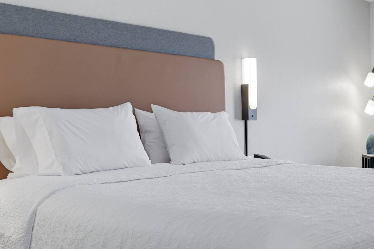 A neatly made bed with white linens, a headboard with a grey and brown panel, and a wall-mounted light fixture.