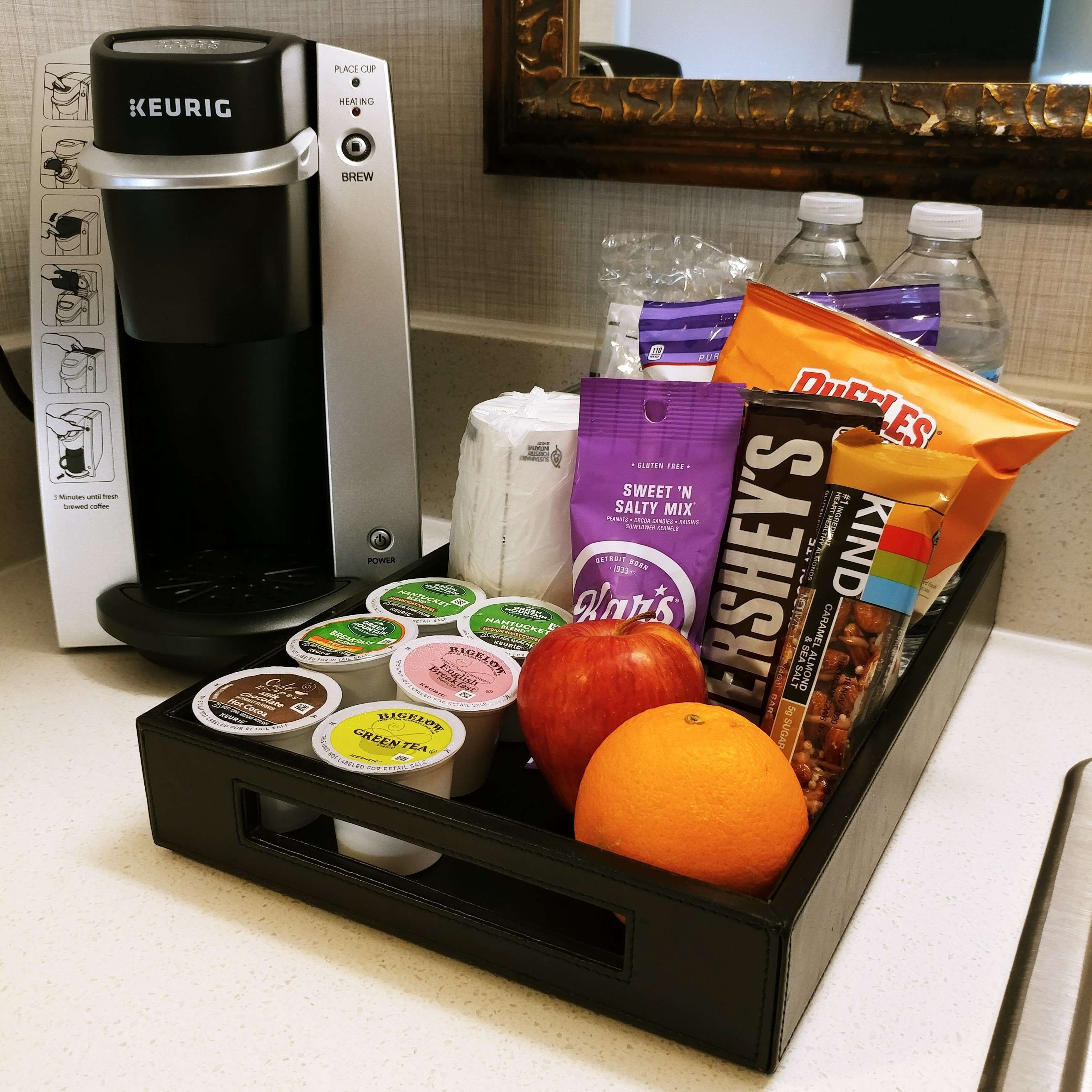 A coffee maker, a tray with coffee pods, a bag of Dufries, a bag of Hershey's, a bottle of water, and a fruit basket.