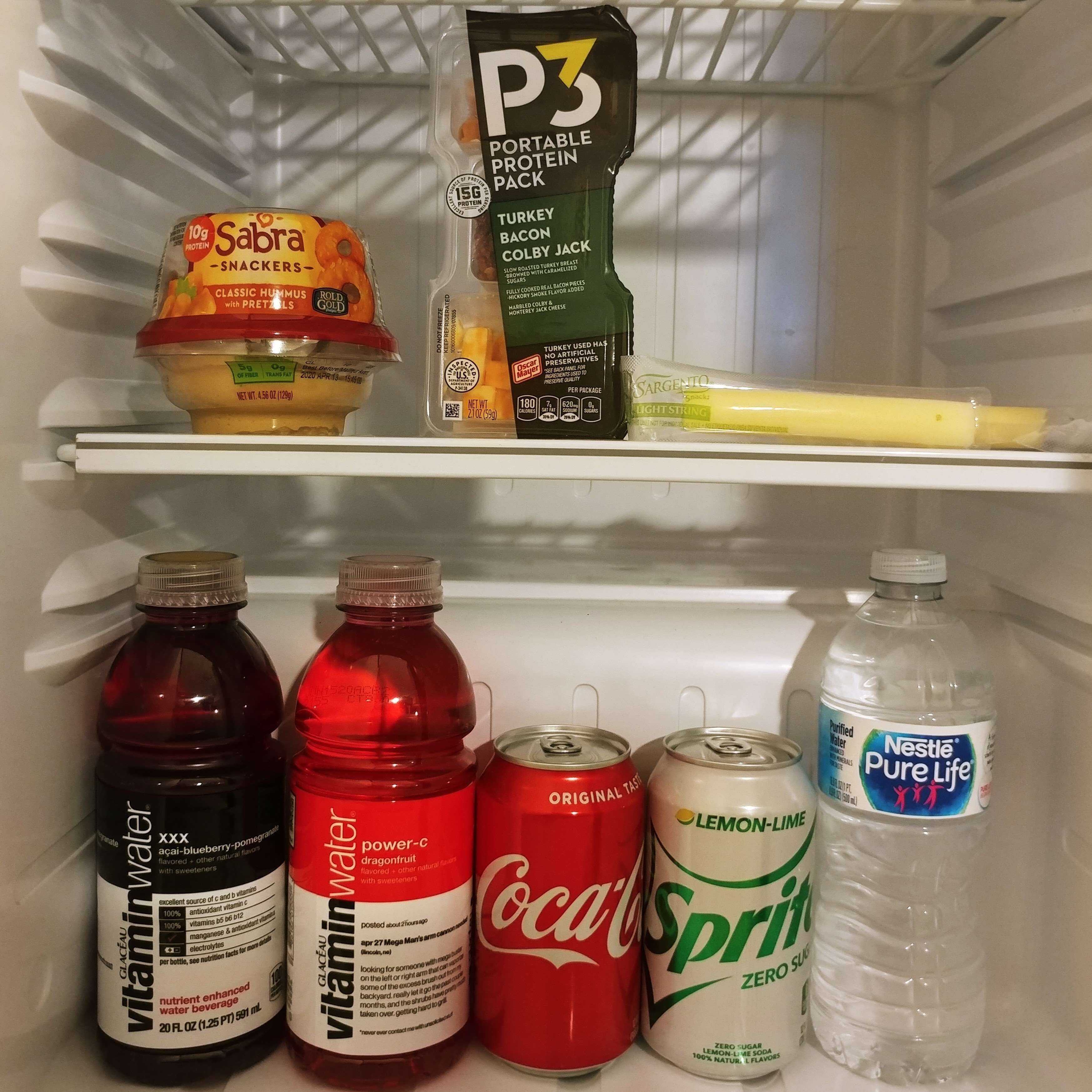 A refrigerator containing various food and drink items including Sabra snacks, a turkey bacon colby jack pack, Sargento light string cheese, vitamin water, Coca-Cola, Sprite, and Nestlé Pure Life water.