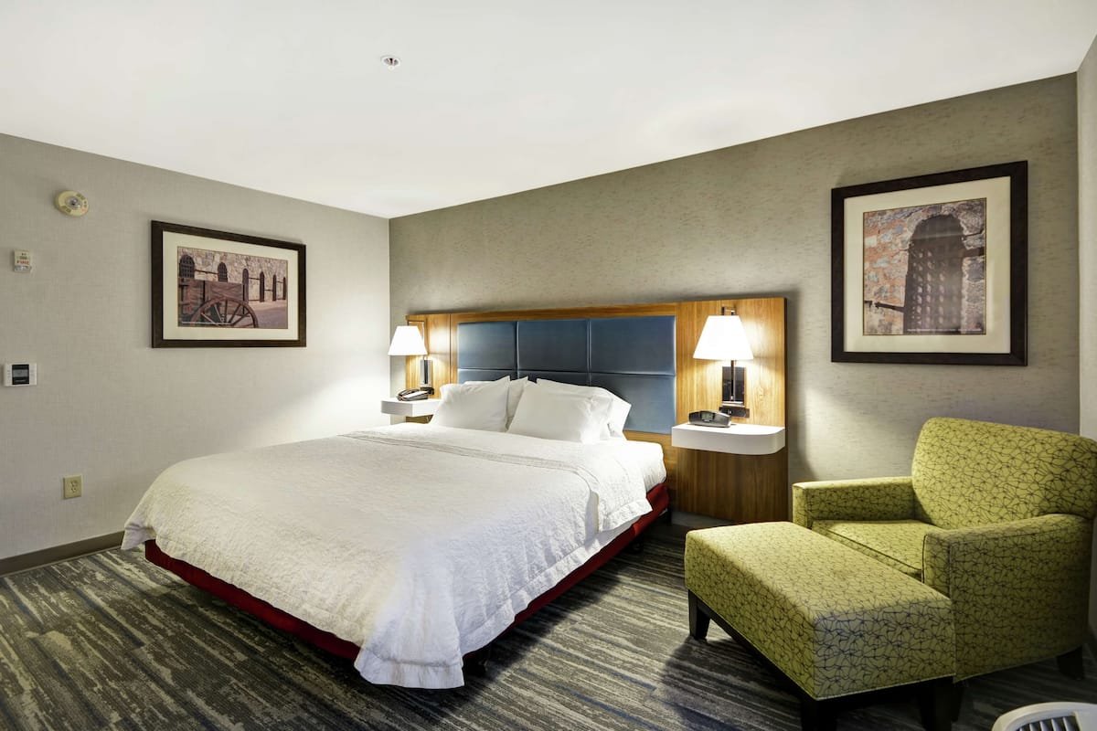 Ein Hotelzimmer mit einem großen Bett, zwei Nachttischen mit Lampen, einem grünen Sessel und zwei gerahmten Bildern an den Wänden.