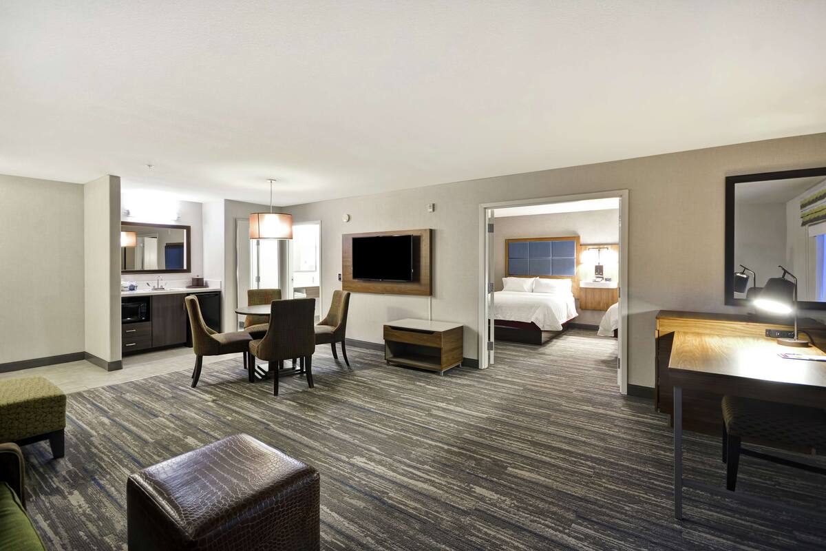 Ein Hotelzimmer mit Küchenzeile, einem an der Wand befestigten Fernseher, einem Bett, einem Schreibtisch mit Lampe und einer Sitzecke mit Stühlen und einem kleinen Tisch.