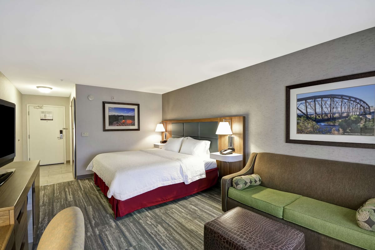 Ein Hotelzimmer mit einem Bett, einem grünen Sofa, einem Schreibtisch mit Fernseher und einem gerahmten Bild einer Brücke.