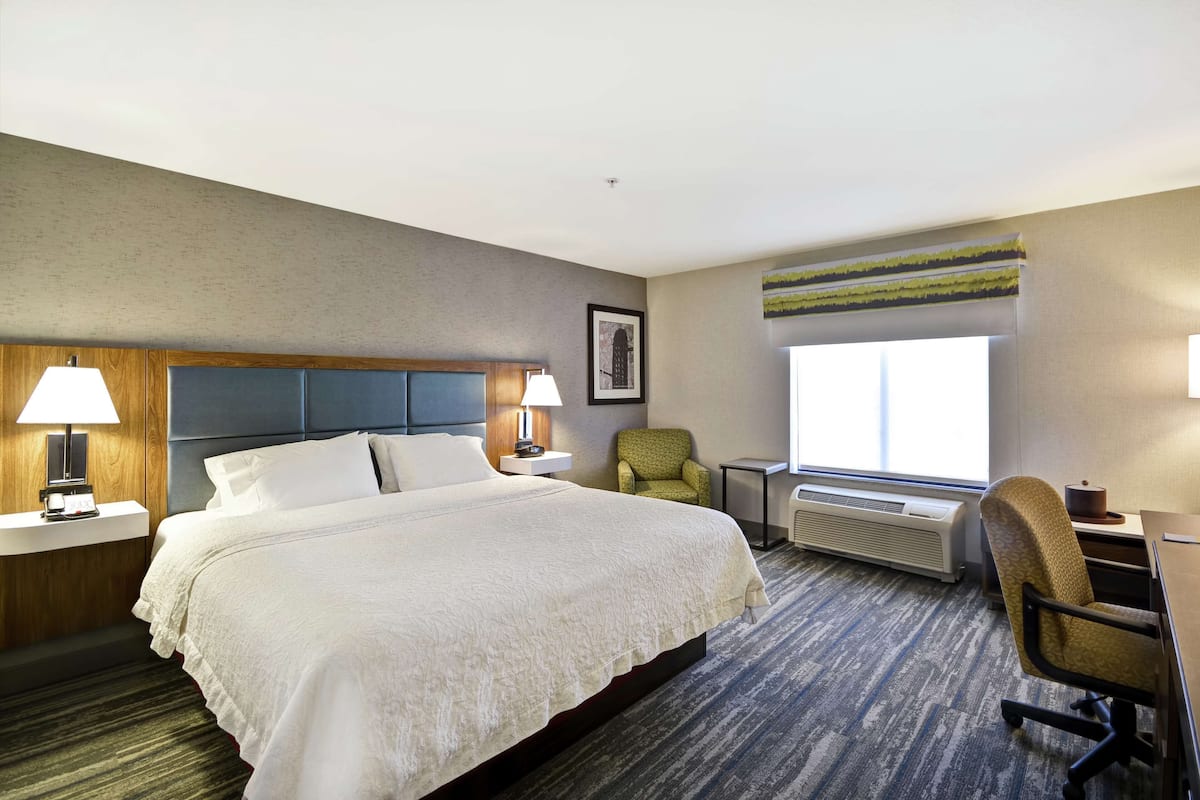 Ein Hotelzimmer mit einem großen Bett, zwei Nachttischlampen, einem Sessel, einem Schreibtisch und einem Fenster mit Blick.