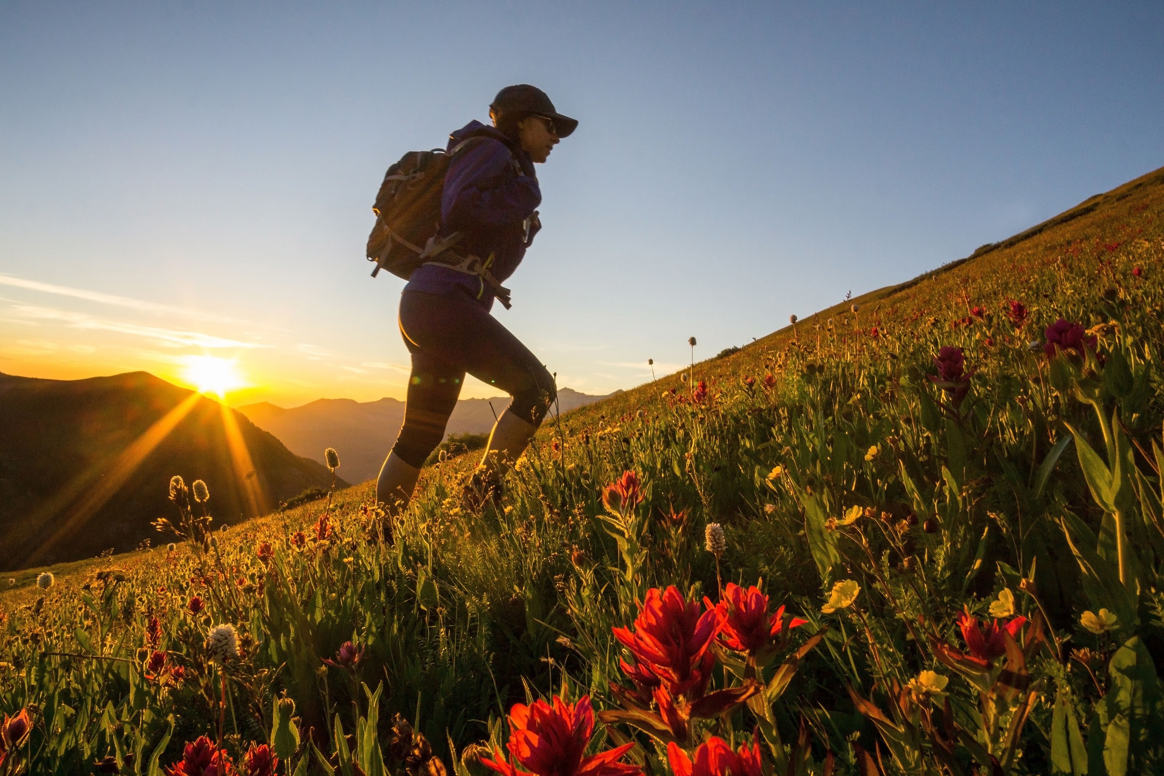 Eine Person wandert mit Rucksack einen grasbewachsenen Hügel hinauf, im Hintergrund sind Berge und die untergehende Sonne zu sehen.