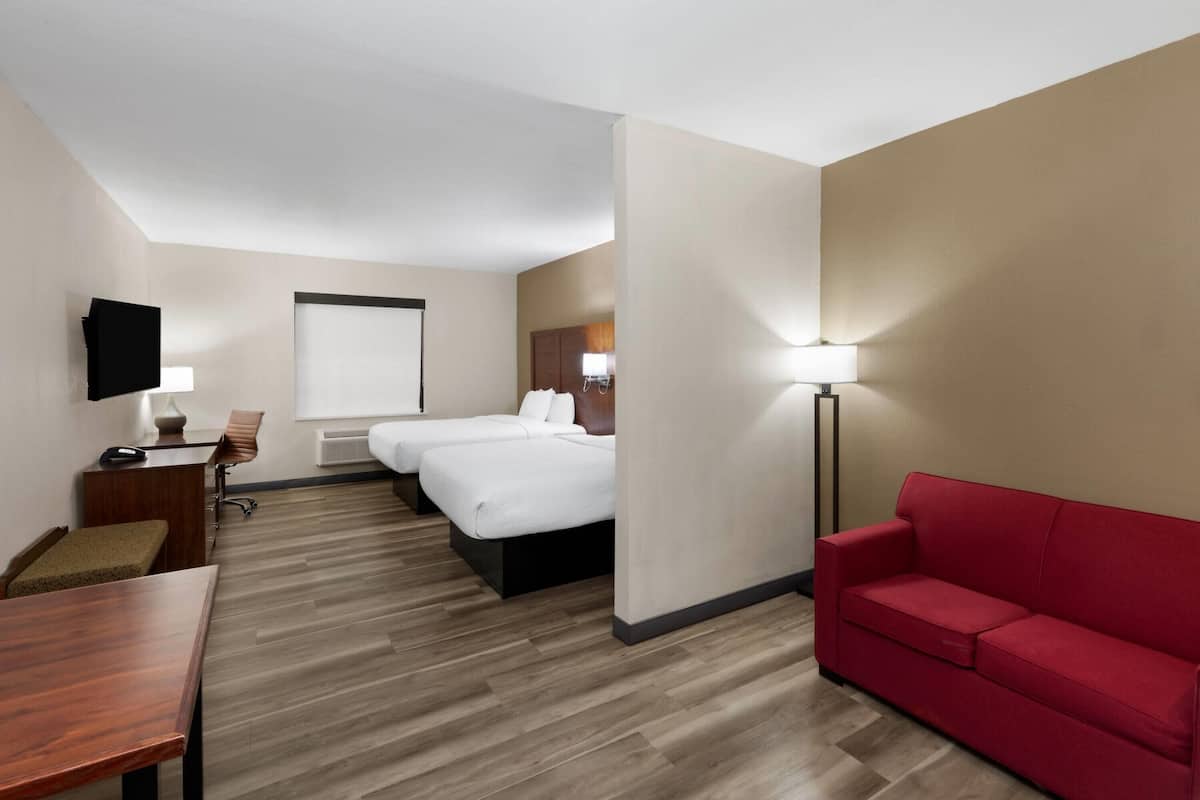 A hotel room with a wooden floor, a red sofa, a desk with a chair, a bed, a TV, and a lamp.