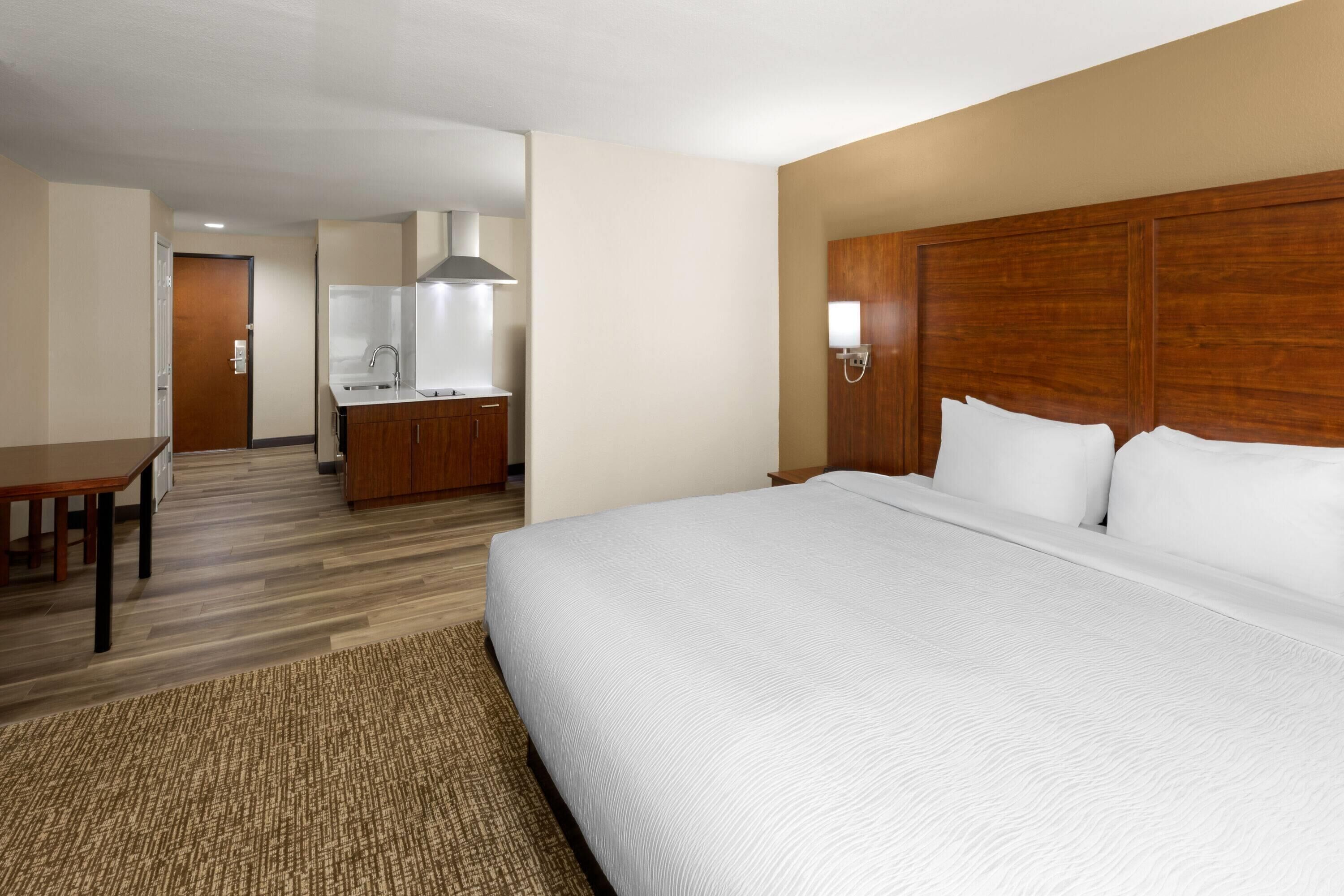 Una habitación de hotel con una cama grande, cabecera de madera, un escritorio pequeño y una kitchenette al fondo.