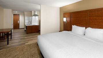 Una habitación de hotel con una cama grande, cabecera de madera, un escritorio pequeño y una kitchenette al fondo.