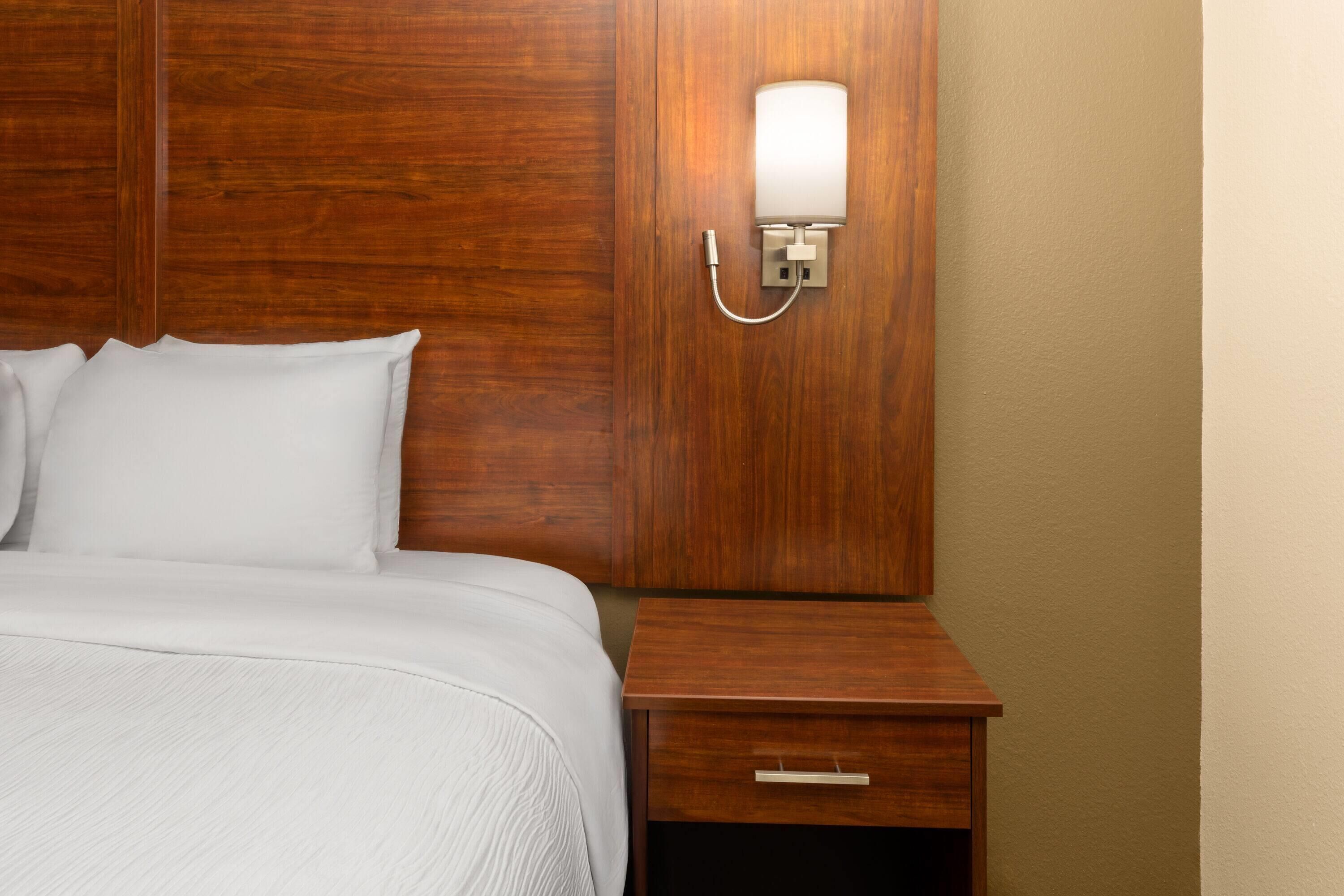 Un lit bien fait, avec du linge de lit blanc, une tête de lit en bois, une table de chevet dotée d’un tiroir et un luminaire fixé au mur.