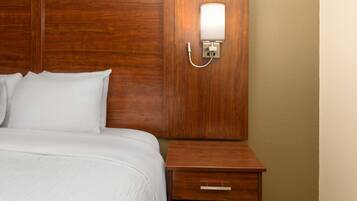 A neatly made bed with white linens, a wooden headboard, a bedside table with a drawer, and a wall-mounted light fixture.