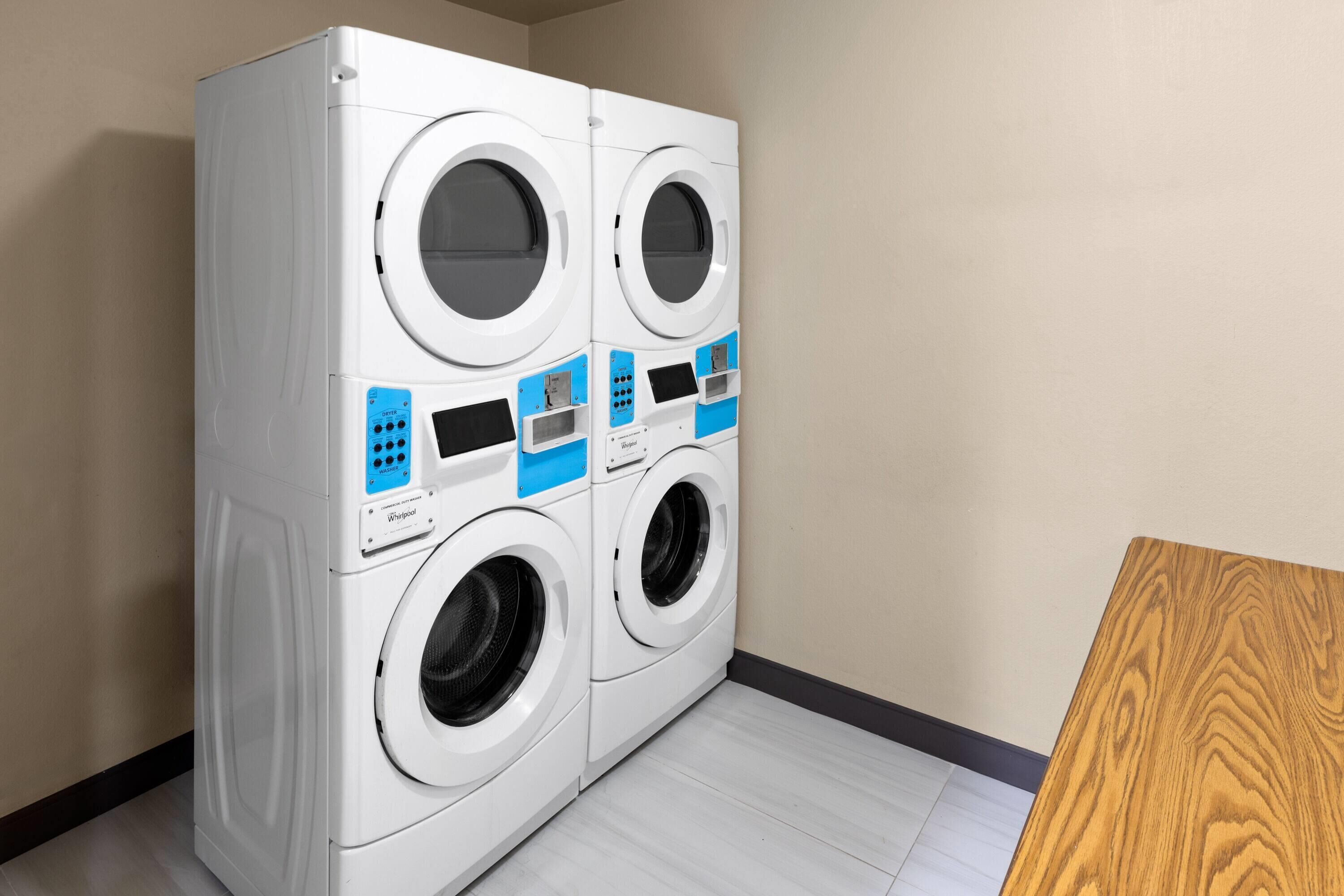 A four-door front-loading washer and dryer unit with control panels, located in a room with light-colored wooden flooring and a plain wall.