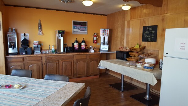 A kitchen with wooden cabinets, a coffee machine, a microwave, and a refrigerator. There is a chalkboard with the text "Food adds the flavor to life..." and a sign that says "MILK AND JUICE".