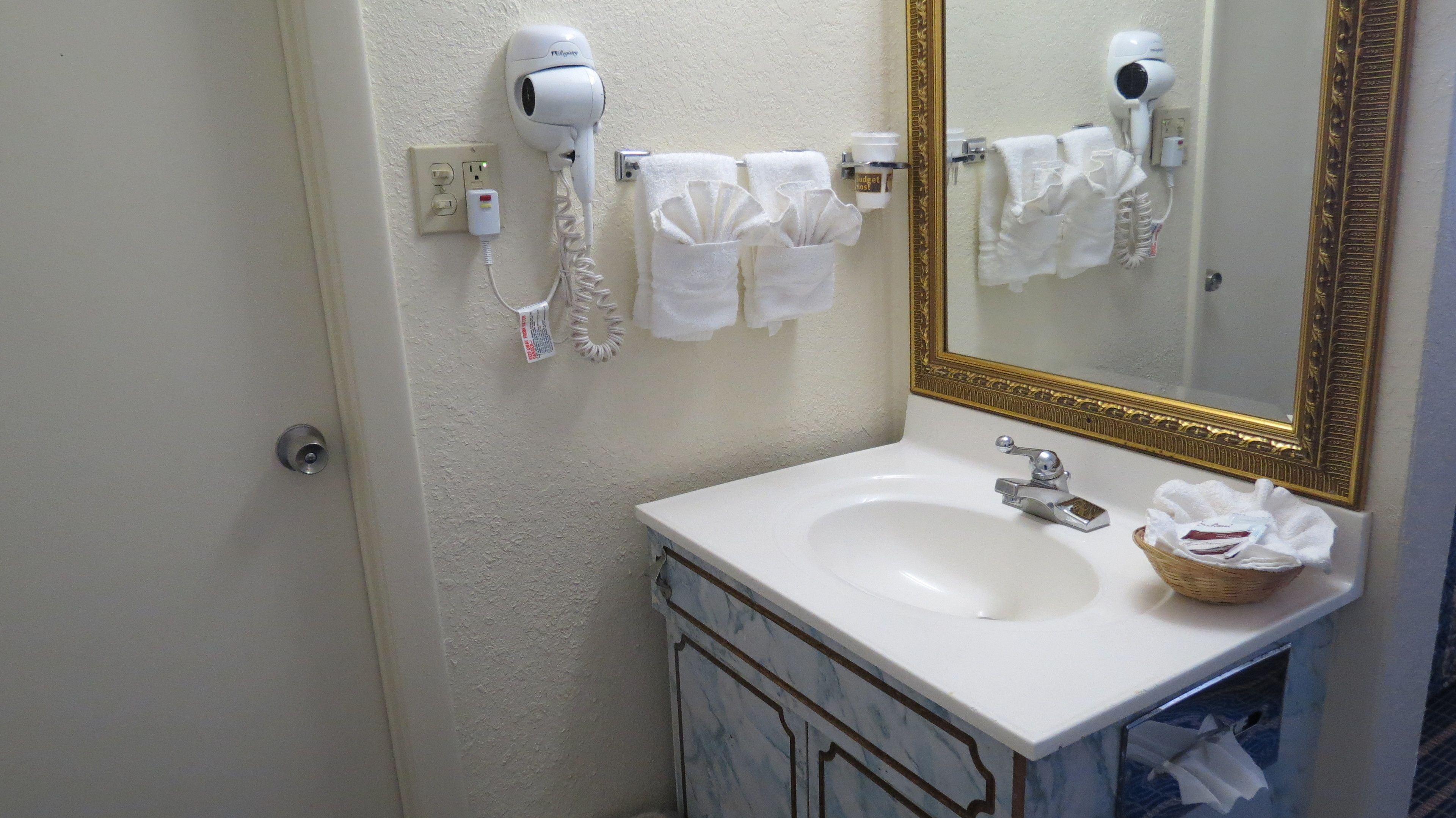 Une salle de bain avec un lavabo blanc, un miroir avec un cadre doré et deux sèche-cheveux fixés au mur.