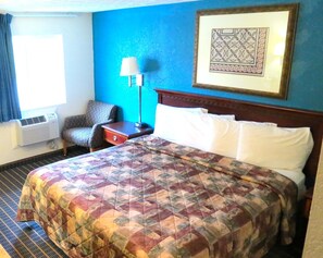 Quarto de hotel com cama grande, uma cadeira, mesa de cabeceira, abajur e quadro na parede.