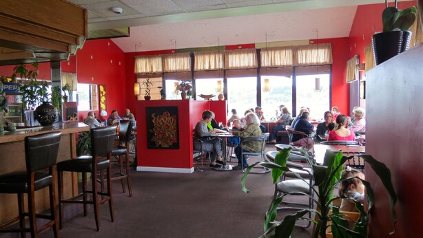 A restaurant with red walls, multiple tables, and chairs. There are potted plants and a large window with curtains.