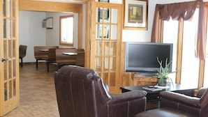 A living room with leather sofas, a flat-screen TV, a dining area with wooden furniture, and a large window with curtains. - Budget Host Sundowner Motor Inn (Kadoka)