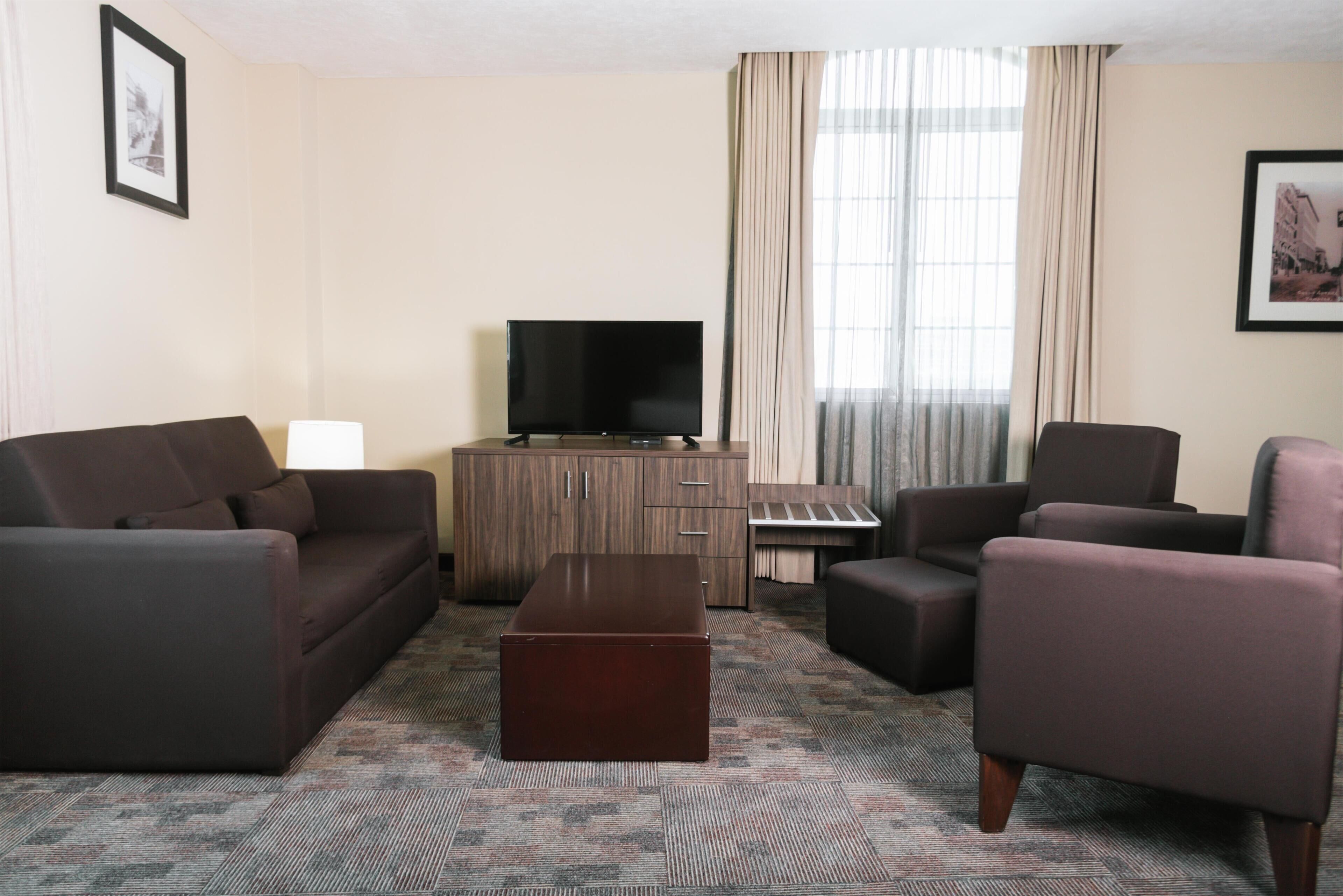 A hotel room with a sofa, armchair, ottoman, TV, cabinet, and curtains.